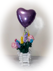 A small white basked made of white balloons is filled with balloons contorted to resemble wild flowers. Floating in the middle of the flowers is a foil lavender heart balloon.