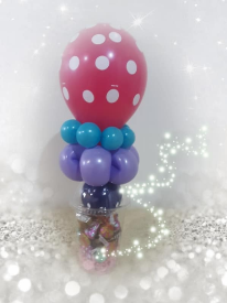 A round pink latex balloon with white polka dots sits atop a lavender latex  balloon contorted into the shape of a flower, surrounded by turquoise beads. The balloons sit on top of a clear domed cup filled with candy.