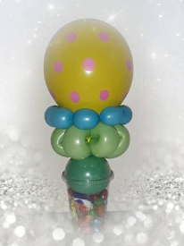 A round yellow latex balloon with pink polka dots sits atop a green latex  balloon contorted into the shape of a flower, surrounded by turquoise beads. The balloons sit on top of a clear domed cup filled with candy.