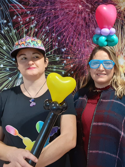 Two women stand next to each other. The first woman wears a baseball hat and stands holding balloon wand. The wand is a yellow heard, on top of a black staff. Next to her is a woman wearing blue sun glasses, and a balloon hat. The balloon hat is a magenta heart on top of a turquoise flowers, surrounded by purple beads.