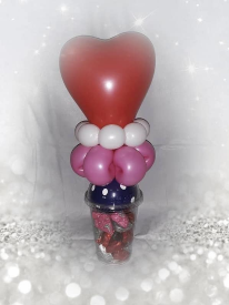 A heart shaped red latex balloon sits atop a magenta latex  balloon contorted into the shape of a flower, surrounded by white beads. The balloons sit on top of a clear domed cup filled with candy.
