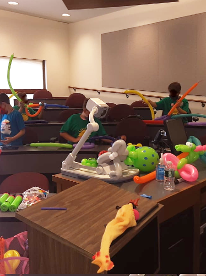 A lecture hall is filled with children who are at  tables making balloon animals. In front of the children is a podium with a yellow and orange rubber chicken on it, and next to that is a table with a  white  projector surrounded by balloon animals.