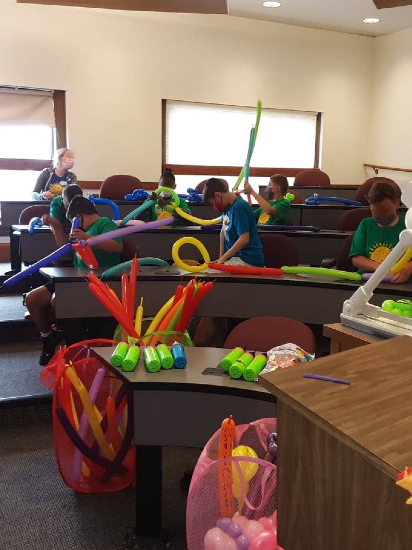 A lecture hall is filled with children who are at  tables making balloon animals. In front of the children is a table filled with green and pink balloon hand pumps, and a basket of multicolored balloons.